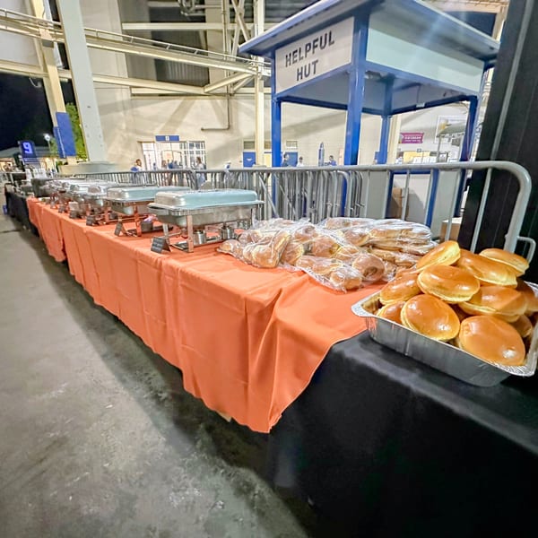A long buffet table with LX BBQ food warmers and trays of buns under a "Helpful Hut" sign.