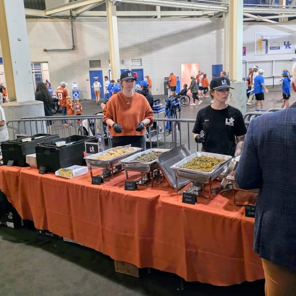 Two LX BBQ staff members serve food from chafing dishes on an orange-covered table at an event.