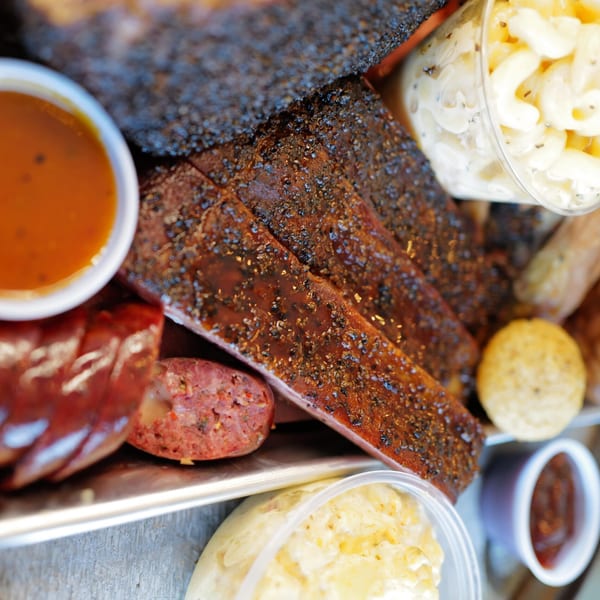 Close-up of LX BBQ ribs, sausage, mac and cheese, and dipping sauces on a metal tray.