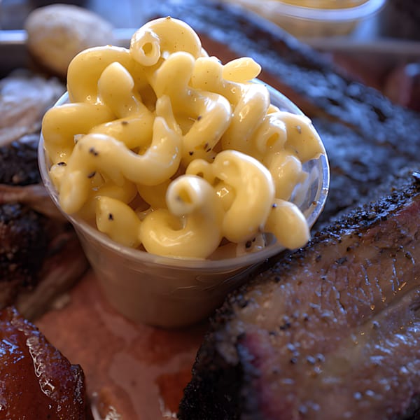 Creamy mac and cheese in a clear cup, surrounded by dark, smoky BBQ brisket and ribs.