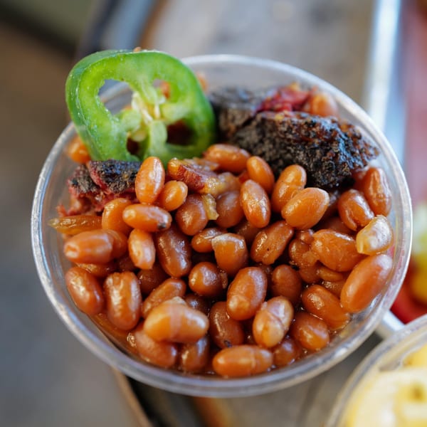 Close-up of LX BBQ baked beans with burnt ends and a jalapeño slice in a clear cup.