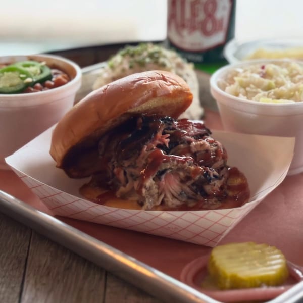 LX BBQ pulled pork sandwich with sides of baked beans, coleslaw, potato salad, and a pickle.